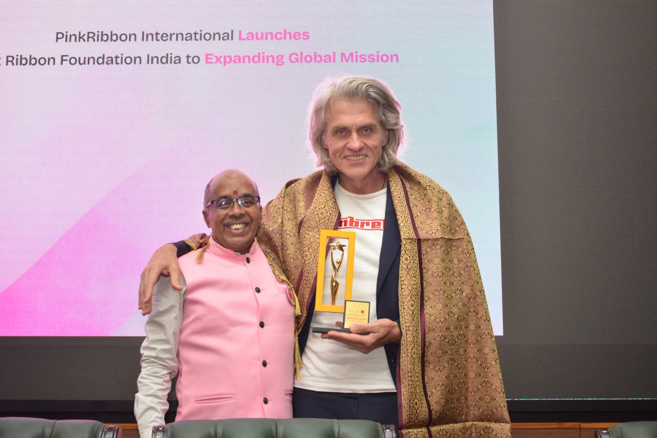 Two launch participants pose on stage with a trophy in front of the PinkRibbon International launches Pink Ribbon Foundation India banner
